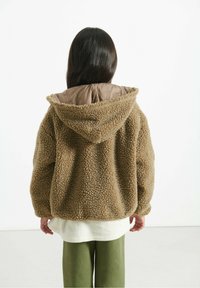Bambino con lunghi capelli scuri che indossa una giacca morbida con cappuccio marrone, una camicia bianca e pantaloni verdi, in piedi di fronte a uno sfondo bianco.