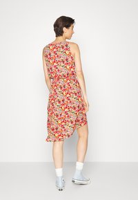 Vestido floral com um padrão vibrante em vermelho, amarelo e verde. Design sem mangas com um hem esvoaçante e cintura ajustada. Usado com ténis de cano alto azuis.