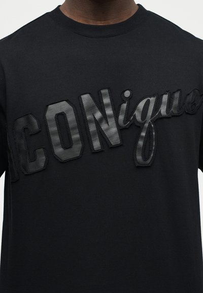Κοντινό πλάνο ενός ατόμου που φοράει μαύρο πουκάμισο με ανάγλυφα, γυαλιστερά μαύρα γράμματα που γράφουν «ICONique» στο στήθος.