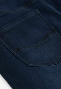 Tejido de mezclilla azul oscuro con una superficie texturizada, que presenta un bolsillo trasero rectangular, delineado con costuras en contraste.