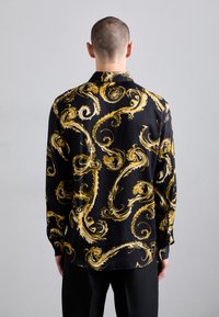 Camicia a maniche lunghe in seta nera con audaci motivi dorati a spirale. Presenta un colletto, dettagli con bottoni e un orlo dritto.