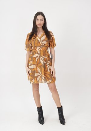 Frau mit langen dunklen Haaren, die ein braun-gelb gemustertes Blumenkleid und schwarze Stiefeletten trägt und vor einem schlichten weißen Hintergrund steht.