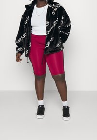 Černá bunda z umělé kožešiny s bílým logem, bílým přiléhavým tričkem, lesklé burgundské cyklistické šortky a černé tenisky s bílými detaily.