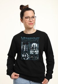 Donna con occhiali che indossa un felpa nera con sei scene e il testo "Mercoledì" dalla serie TV Mercoledì.
