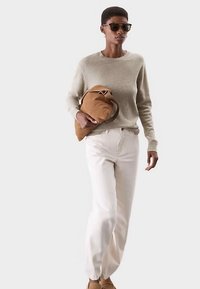 Jasnobeżowy sweter, białe dżinsy i brązowa torba. Dodatki to okulary przeciwsłoneczne. Model stoi pewnie na tle o neutralnym kolorze.