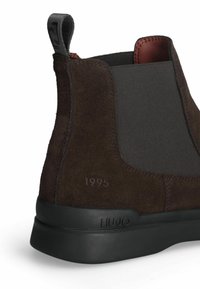 Stivaletto in suede marrone con lati elasticizzati neri, suola in gomma e linguetta. Presenta la scritta incisa "est 1995" sul lato e il logo del marchio sulla suola.