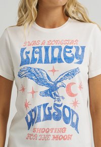 Balta kokvilnas T-kreklā ar zilu un rozā grafisko tekstu un ērgļa dizainu, kurā ir zvaigznes un pusmēness. Apaļš kakla izgriezums, īsas piedurknes.