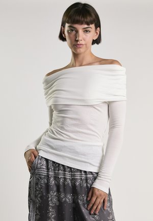 Młoda kobieta o krótkich włosach, ubrana w białą bluzkę odkrywającą ramiona i czarno-białe spodnie w wzory, pozująca z jedną ręką w kieszeni.