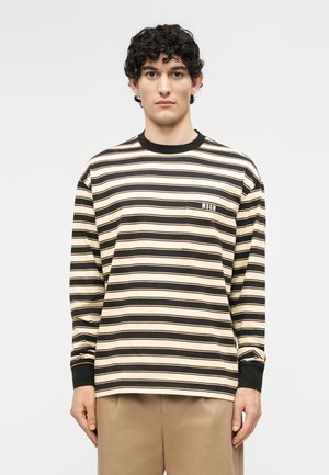 Homme aux cheveux bouclés et foncés portant un t-shirt à manches longues beige et noir à rayures horizontales avec un patch "MSGM", debout devant un fond uni.