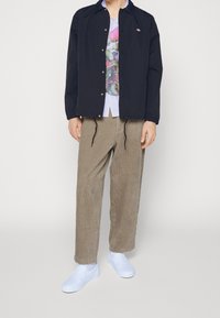 Black lightweight jacket over a multicolored floral graphic T-shirt, paired with beige corduroy pants and white slip-on shoes.