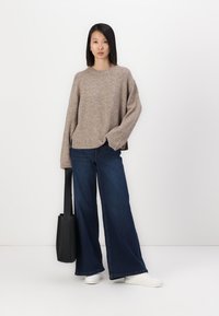 Beige stickad tröja med långa ärmar, parat med vida mörkblå jeans. En svart totebag hänger över axeln, och vita sneakers är på fötterna.