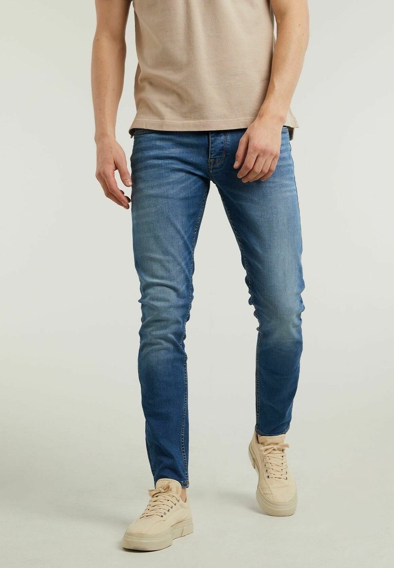 Jean skinny bleu délavé, avec une finition usée, présentant un design classique à cinq poches et une texture usée, associé à des baskets beige clair.