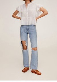 Biała, krótka, zapinana na guziki bluzka z plisowanymi detalami, zestawiona z jasnoniebieskimi, postrzępionymi dżinsami z dużymi przetarciami oraz brązowymi sandałami na paski.
