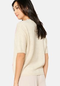 Cremfarbener, gestrickter Pullover mit kurzen Ärmeln, der eine weiche Textur und einen lockeren Schnitt aufweist, von hinten betrachtet. Keine sichtbaren Muster oder Akzente.