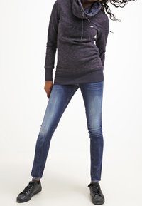 Žena v tmavě šedé mikině s kapucí, modrých skinny džínách a černých teniskách, stojící s nohama rozkročenýma na bílém pozadí.