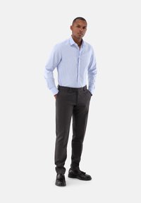 Helles blau gestreiftes Hemd mit langen Ärmeln, kombiniert mit dunkelgrauen, maßgeschneiderten Hosen und schwarzen Schuhen, stehend mit Händen in den Taschen.