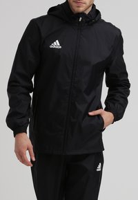 Mies, joka on pukeutunut mustaan Adidas-huppariin ja siihen sopiviin housuihin, seisoo yhdellä kädellä takin taskussa yksinkertaista taustaa vasten.