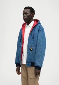 Τζάκετ από τζιν με κόκκινη επένδυση από πλισέ, κουκούλα με κορδόνι, μπροστινή τσέπη και μαύρους ριμπ μανίκους. Διαθέτει λογότυπο στη θήκη του στήθους.