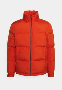 Ljust orange pufferjacka med hög krage, framdragkedja, sido fickor och små logotyp-patchar på bröstet och vänster ärm.