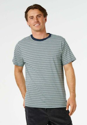 Jeune homme souriant, portant un T-shirt blanc à manches courtes avec de fines rayures horizontales noires et un pantalon foncé, debout devant un fond uni.