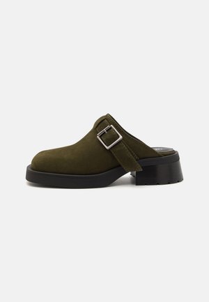 Olive rohelise suede'ist mule madala, robustse musta kontsaga. Omab külgrihma hõbedase pandlaga reguleerimiseks. Üldine tekstuur on sile.