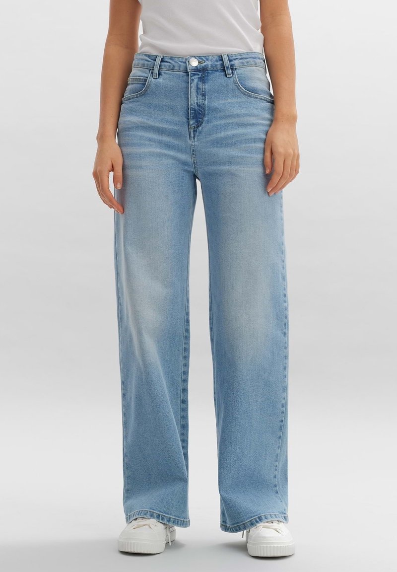 Personne portant un jean taille haute large bleu clair, associé à des baskets blanches et une chemise blanche rentrée, debout devant un fond uni.
