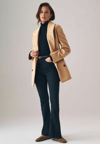 Beige Wollmischung-Blazer über einem schwarzen Rollkragenpullover, kombiniert mit dunklen Schlagjeans und braunen Ballerinas. Glatte Textur, taillierte Passform und lässiger Stil.