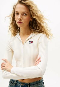 Felpa bianca con zip, a coste, con maniche lunghe e design aderente. Presenta un patch del logo Tommy Jeans sul lato sinistro del petto.