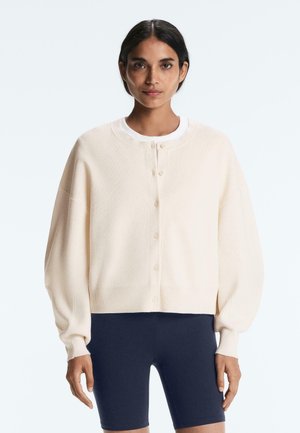 Kobieta o ciemnych włosach ubrana w kremowy kardigan na guziki, nałożony na białą koszulę, oraz granatowe dopasowane szorty, stojąca na jasnym tle.