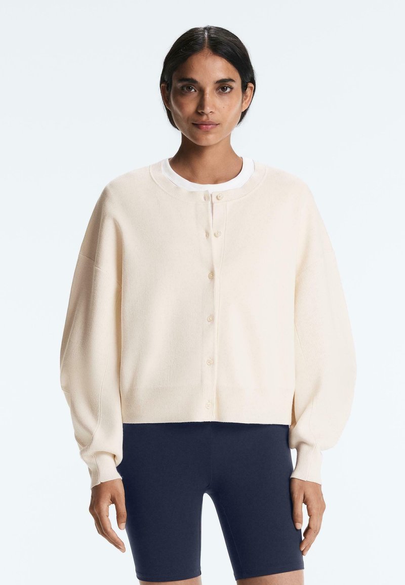 Femme aux cheveux foncés portant un cardigan crème boutonné sur une chemise blanche et un short bleu marine ajusté, debout devant un fond clair.