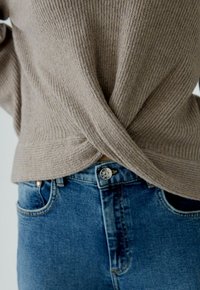 Pull en beige côtelé avec un design de dos torsadé, associé à un jean bleu taille haute avec un bouton en métal et une texture délavée.