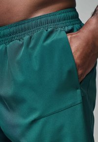 Grønne atletiske shorts lavet af glat stof, med en elastisk talje og sidelomme. Materialet har en diskret glans og blød tekstur.