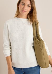 Donna che indossa un maglione a maglia bianco con macchie e jeans blu, mentre porta una borsa a spalla imbottita di colore verde oliva su uno sfondo beige uniforme.