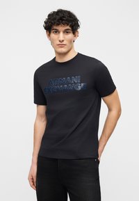 Νεαρός άνδρας με σγουρά μαλλιά, φοράει μαύρο μπλουζάκι Armani Exchange και μαύρο τζιν, στέκεται μπροστά σε ένα απλό φωτεινό φόντο.