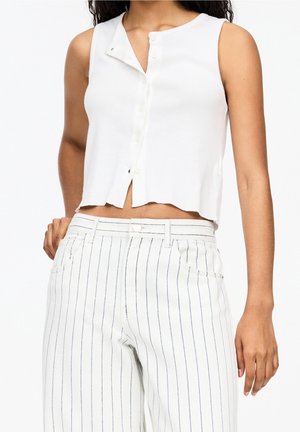 Crop top blanc sans manches boutonné, associé à un pantalon blanc avec de fines rayures verticales noires.