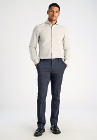 Hellgraues Button-down-Hemd mit einem subtilen Muster, kombiniert mit dunkelblauen, flachen Hose. Das Model steht selbstbewusst mit den Händen in den Taschen.