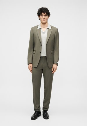 Młody mężczyzna o kręconych włosach, ubrany w oliwkowo-zielony garnitur, jasnoszary sweter z dekoltem w serek oraz czarne eleganckie buty, stojący na tle jednolitego tła.