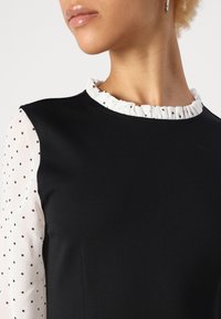 Abito nero con un colletto bianco arricciato e maniche trasparenti a pois. Il tessuto è liscio con un design aderente, evidenziando un contrasto chic.