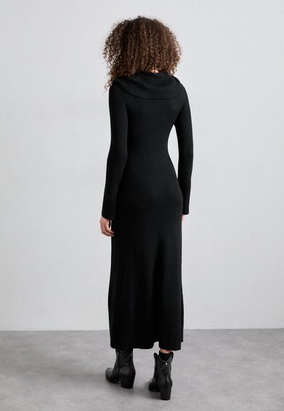 Black ribbed long-sleeve dress with a cowl neckline, form-fitting silhouette, and a flared hem, worn with black heeled ankle boots.