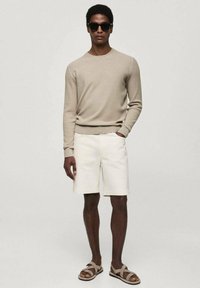 Beige ribbed knit sweater, white cotton shorts, and brown sandals. Model wearing dark sunglasses, standing against a neutral background.