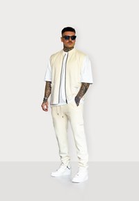 Beige Zip-Weste über einem weißen T-Shirt, passende beige Jogginghosen und weiße Sneaker. Hat Taschen und ein sportliches Design.