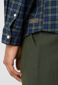Kariertes Hemd in blau-grüner Baumwolle, mit geknöpftem Manschette und einem Etikett mit "SCOTCH & SODA"-Details. Olivgrüne Hose mit Seitentasche.