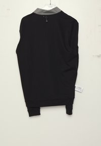 Chemise noire à manches longues avec col gris suspendue sur un cintre transparent sur fond blanc, montrant la vue arrière et l'étiquette du vêtement.
