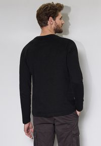 Black long-sleeve t-shirt made of cotton, featuring a round neck and fitted sleeves. Worn with dark cargo pants, viewed from the back.
