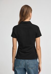 Camisa polo negra de manga corta con cuello plano, confeccionada en tejido suave. La vista trasera muestra un sutil entalle y un dobladillo limpio.