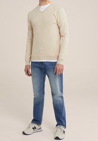 Beige v-ringad tröja med långärmar, kombinerad med ljusblå jeans. Modellen bär vit t-shirt under och grå sneakers. Enkel design.
