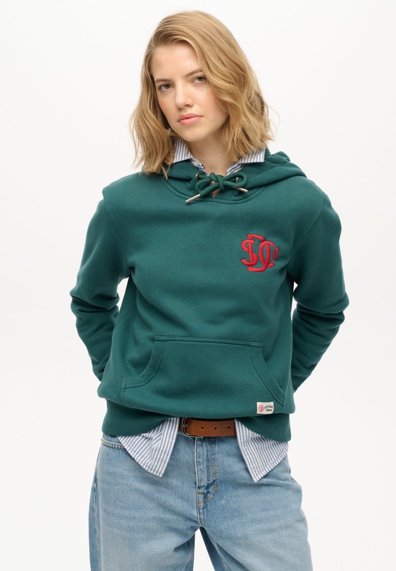 Groen hoodie met een voorzak, voorzien van een rood geborduurd logo, trekkoord bij de nek, gedragen over een blauw-wit gestreept shirt.