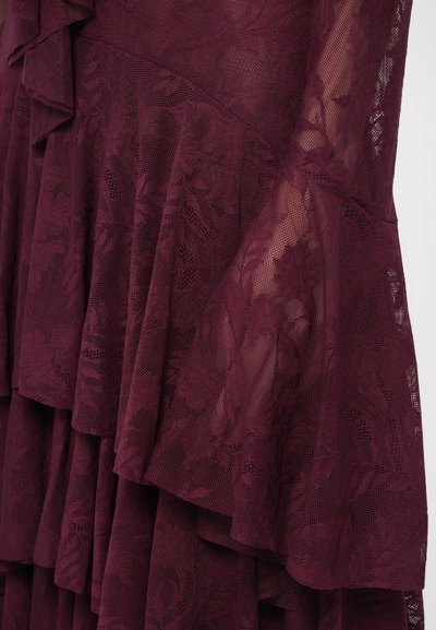 Abito in pizzo color bordeaux con balze sovrapposte, motivi floreali e tessuto trasparente. Maniche svasate che aggiungono dimensione al design.