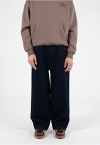 Sudadera marrón con un bolsillo frontal, combinada con jeans vaqueros de pierna ancha en tono oscuro. Los jeans presentan costuras visibles y una textura ligeramente desgastada.