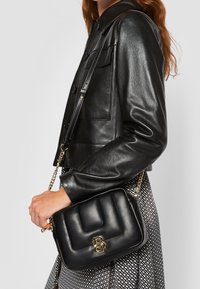 Černá kožená crossbody taška s prošívaným vzorem, zlatými kovovými detaily a řetízkem; vystavená s rukou, která ji drží, na pozadí tištěné sukně.